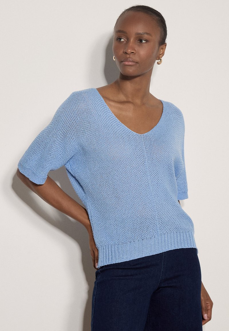 Femme portant un pull léger à manches courtes en tricot bleu clair avec col en V et pantalon bleu foncé, debout avec une main sur la hanche devant un fond uni.