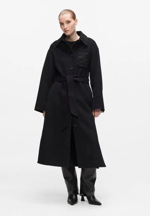 Jeune femme aux cheveux plaqués en arrière portant un long manteau noir ceinturé sur un pantalon noir et des bottes noires à bout pointu.