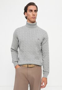 Grau kabelgestrickter Rollkragenpullover mit gerippten Bündchen, ausgestattet mit einem kleinen Logo auf der Brust, kombiniert mit hellbraunen Hosen und einem Gürtel.