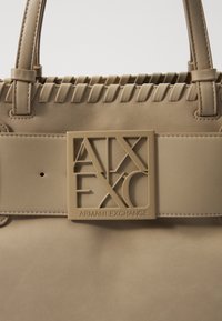 Beige leather handbag with a structured design. Features woven detailing and a prominent tan logo plaque on a wide strap. Smooth texture.