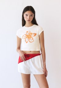 Witte cropped t-shirt met oranje bloemenontwerp, gecombineerd met witte shorts en een rode taillebandaccent. Zachte textuur, casual ontwerp.