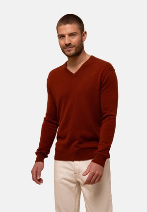 Rdzawy sweter w serek z miękkiej, teksturowanej dzianiny. Dopasowany krój z prążkowanymi mankietami i dołem. Noszony na jasnych dżinsach.