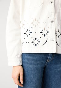 Valge denim jakk, mille alläär on lillelise lõikega, koos teksadega ja külje peal nähtava käega. Esiküljel on nupud.