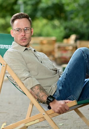 Holzliegestuhl mit grünem Sitz aus Segeltuch. Person in einem hellbeigen Hemd und blauen Jeans, die eine Holzarmlehne hält, mit sichtbarem Tattoo.