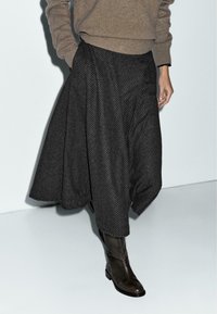 Kvinde iført en beige sweater, sort ternet midiskørt og mørkebrune læderstøvler, stående mod en ensfarvet lys baggrund.