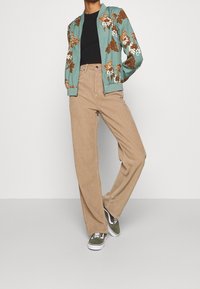 Chaqueta bomber verde azulado con patrón floral, camiseta negra ajustada, pantalones de pana beige de pierna ancha y zapatillas verdes con suelas blancas.