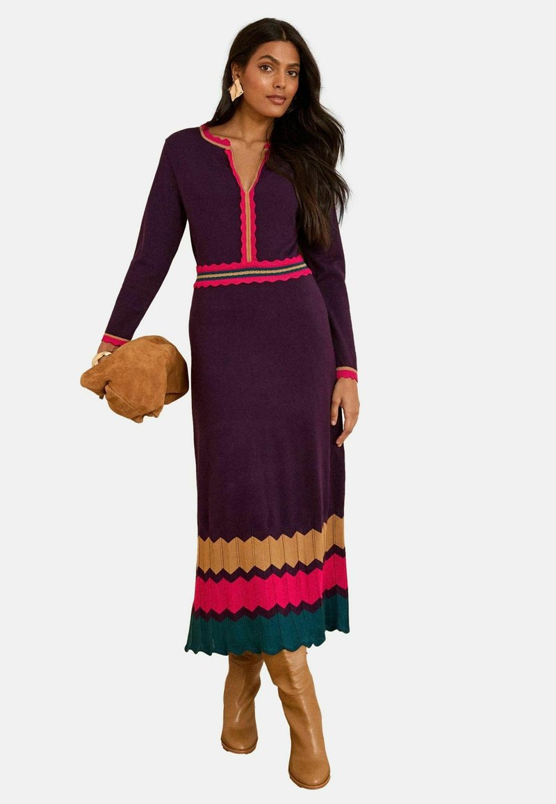 Abito lungo e aderente viola con scollatura a V, bordi ondulati e orlo zigzag colorato in rosa, verde e oro. Indossato con stivali color beige fino al ginocchio.