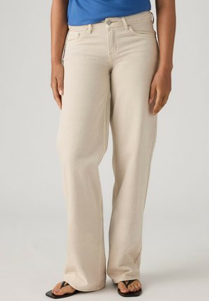Personne portant un pantalon beige à jambes larges, un haut bleu et des tongs noires, debout devant un fond clair uni.