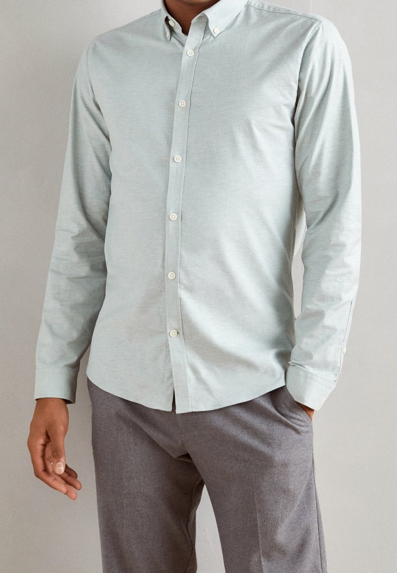 Jasnozielona koszula na guziki z długimi rękawami, miękka w dotyku, z kołnierzykiem i białymi guzikami, zestawiona z szarymi spodniami.