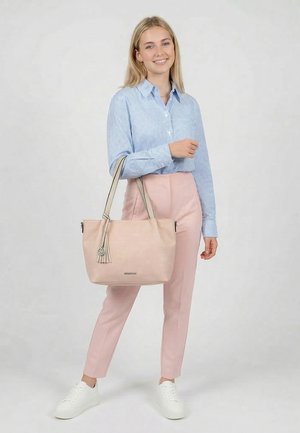 Jonge vrouw lacht, draagt een lichtblauw geruit overhemd, roze broek, witte sneakers, houdt een grote lichtroze totebag met kwastdetail vast.