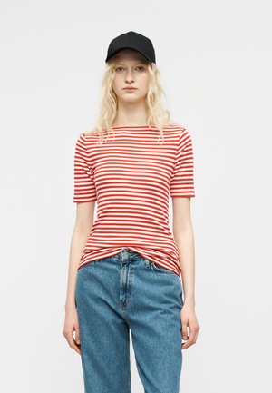 Ung kvinde med langt blondt hår iført en sort kasket, rød- og hvidstribet kortærmet trøje og blå jeans, stående mod en ensfarvet baggrund.