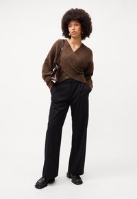 Brązowy sweter w miękkim dzianinowym splotach, połączony z czarnymi spodniami w szerokie nogawki. Modelka ma przy sobie małą ciemną torebkę i nosi czarne loafersy.