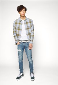 Camisa de cuadros en azul y verde olivo; camiseta blanca debajo. Jeans claros rasgados y zapatillas verdes con suelas blancas. Fondo neutro.