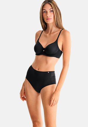 Vrouw staand, gekleed in een zwarte beugelbeha met dunne bandjes en bijpassende hooggetailleerde onderbroek, tegen een effen lichte achtergrond.