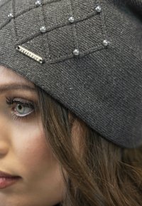 Gray knitted beanie featuring a quilted pattern accented with silver beads and a metallic logo tag, offering a textured appearance.