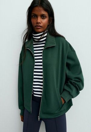 Une femme porte une veste ouverte vert foncé à fermeture éclair sur un col roulé à rayures noires et blanches et un pantalon foncé, devant un mur uni.