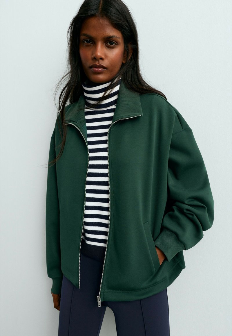 Une femme porte une veste ouverte vert foncé à fermeture éclair sur un col roulé à rayures noires et blanches et un pantalon foncé, devant un mur uni.