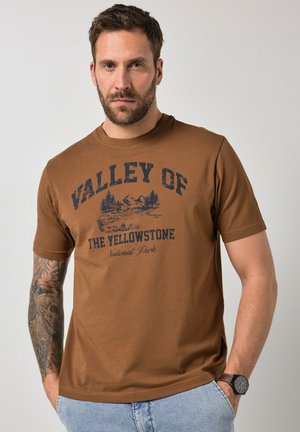 Mann mit kurzen Haaren und Bart, trägt ein braunes T-Shirt mit der Aufschrift „Valley of the Yellowstone National Park“ und hellblaue Jeans, ein Arm tätowiert.