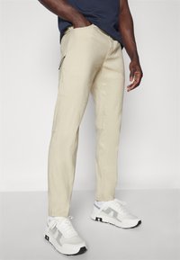 Beige slimfitbyxor med en mjuk textur, med en sidoficka med dragkedja. Matchade med vita och grå sneakers, stående på en neutral bakgrund.