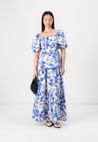 Ensemble floral à deux pièces comprenant un top à manches bouffantes et une jupe maxi, en tissu blanc avec des imprimés botaniques bleus, complété par un sac à main noir.