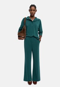 Zielony komplet wypoczynkowy składający się z bluzki z długim rękawem, kołnierzykiem i zapięciem na zatrzaski, zestawiony z spodniami o szerokim kroju. Brązowa, zamszowa torebka na ramię i obuwie.