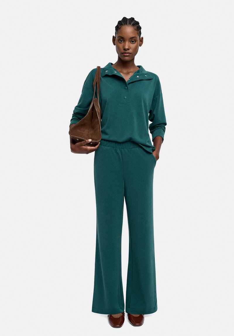 Zielony komplet wypoczynkowy składający się z bluzki z długim rękawem, kołnierzykiem i zapięciem na zatrzaski, zestawiony z spodniami o szerokim kroju. Brązowa, zamszowa torebka na ramię i obuwie.