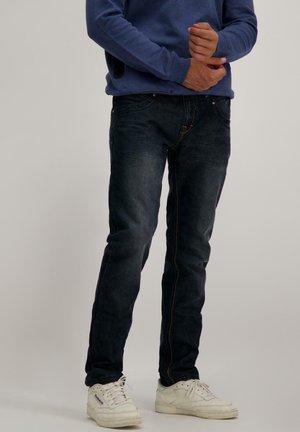 Donkerblauwe slim-fit jeans met contrasterende gele stiksels, gedragen met een blauwe sweatshirt en witte sneakers, waarmee een casual ontwerp wordt getoond.