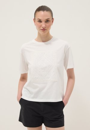 Joven mujer con el pelo recogido, vistiendo una camisa blanca de manga corta con diseño cuadrado texturizado y pantalones cortos negros, manos en los bolsillos, fondo neutro.