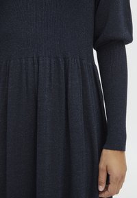 Abito in maglia blu scuro con corpetto testurizzato e gonna svasata. Maniche lunghe, leggera lucentezza e dettaglio di vita arricciata.