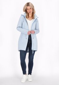 Moteris vilkinti šviesiai mėlyną su kapuco paltą, baltą marškinėlį, tamsias liesas džinsus ir baltus sportbačius, stovinti prieš paprastą baltą foną.