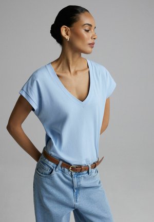 Kobieta z włosami związanymi w kok, ubrana w jasnoniebieską koszulkę z dekoltem w serek, jasne dżinsy oraz brązowy pasek, stojąca na jednolitym szarym tle.