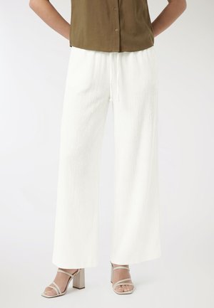 Donna indossa pantaloni bianchi a gamba larga e texture, sandali beige con tacco alto e una camicia verde oliva con bottoni, con le mani dietro la schiena.