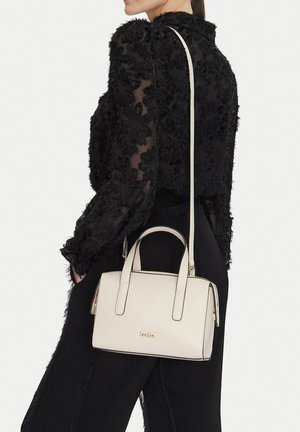 Mujer con blusa y pantalones negros texturizados, llevando un bolso beige rectangular con asas superiores y una correa larga para el hombro.