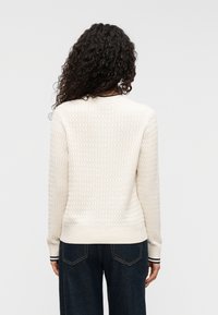 Kreminės spalvos megztinis su tekstūruotu deimanto raštu, ribbed rankogaliais ir vienspalviu juosmeniu su kontrastingomis juostelėmis.