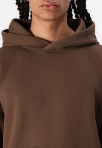 Casaco de capuz castanho com um tecido macio e texturado, apresentando um capuz espaçoso e um corte descontraído. Design minimalista, sem padrões ou acessórios visíveis.
