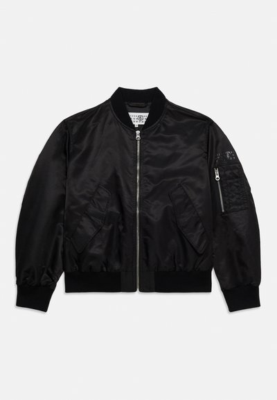 Μαύρο μοδάτο μπουφάν με μπροστινό φερμουάρ, δύο λοξές τσέπες με καπάκια, ριμπ μανσέτες και κάτω μέρος, και τσέπη με φερμουάρ στο αριστερό μανίκι.