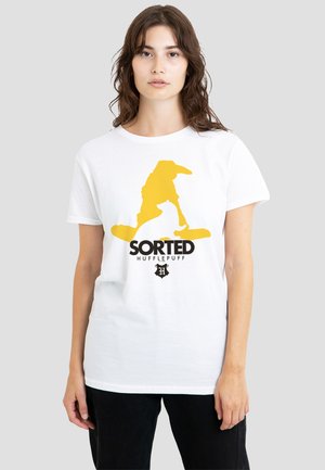 Woman wearing a white T-shirt with yellow silhouette of a wizard and black text "SORTED HUFFLEPUFF" on front, standing against gray background.