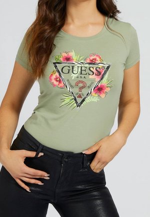 Vrouw draagt een lichtgroen Guess T-shirt met bloemmotief en zwarte broek, poseert met handen in de buurt van de taille.