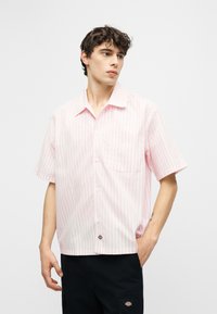 Νέος άνδρας που κοιτάζει δεξιά, φοράει ένα κοντομάνικο πουκάμισο με ρίγες σε ανοιχτό ροζ και λευκό και μαύρο παντελόνι με λογότυπα.