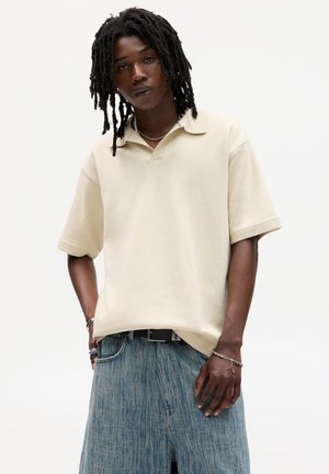 Polo shirt beige a maniche corte realizzato in tessuto morbido, con colletto classico e vestibilità ampia, indossato con pantaloni corti in denim azzurro.