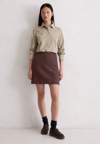 Lichtbeige blouse met kraag, gecombineerd met een getailleerde bordeauxrode rok. Het model draagt bruine instap schoenen en zwarte geribbelde sokken.