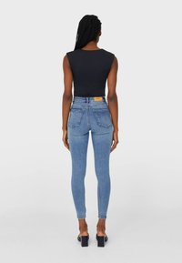 Siniset korkeavyötäröiset skinny farkut, joissa on kaksi takataskua, yhdistetty mustaan ilmanhihaiseen paita. Sileä pinta ja tiukka muotoilu, jossa on vaalea brändilogo.