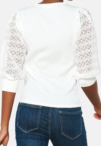 Biały sweter z teksturowanym korpusem i przezroczystymi, koronkowymi bufkami. Dopasowane mankiety i okrągły dekolt. Zestawiony z ciemnoniebieskimi dżinsami.