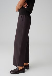 Pantaloni a gamba larga in tessuto marrone scuro, con tasche laterali e una texture morbida, abbinati a scarpe in pelle marrone.
