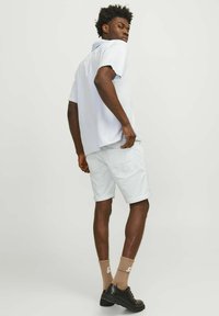 Camisa de manga corta azul claro con corte recto, combinada con pantalones cortos blancos. El modelo lleva calcetines beige y zapatos negros, con un peinado texturizado.
