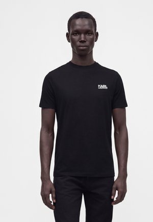 Uomo che indossa una T-shirt nera semplice con il logo "Karl Lagerfeld" sul petto in alto a sinistra, in piedi davanti a uno sfondo bianco semplice.