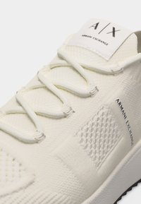 Κοντινό πλάνο σε αθλητικό παπούτσι Armani Exchange κρεμ χρώματος με υφαντό ύφασμα υφής και ετικέτες με το λογότυπο στη γλώσσα και στην πλευρά.