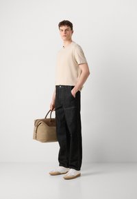 Haut beige à manches courtes, pantalon en denim sombre, baskets blanches avec des accents beiges, tenant un sac de voyage beige avec des accents marron. Design simple et moderne.