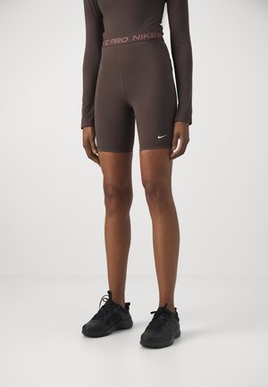 Person iført brune Nike Pro cykelshorts og matchende langærmet top med sorte sportssko, stående mod en hvid baggrund.