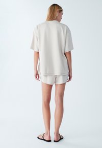 Svetlo bež velika majica s kratkimi rokavi in ravnim robom, združena z usklajenimi kratkimi hlačami. Gladek vzorec, minimalen dizajn in sproščen kroj.